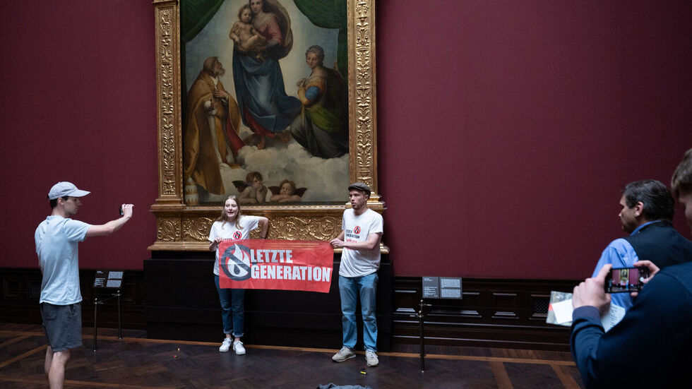 Zwei Mitglieder der Gruppe „Letzte Generation“ stehen mit einem Banner am Gemälde „Sixtinische Madonna“ von Raffael. Beide haben eine Hand am Rahmen festgeklebt. 