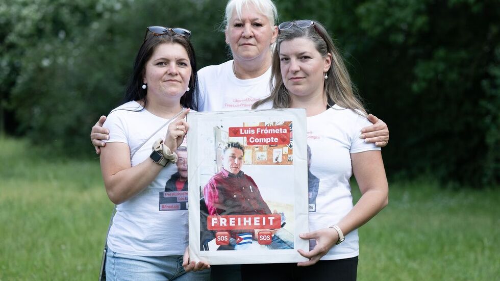 Maria (l) und Janie (r) Frometa Compte, Töchter des inhaftierten Deutsch-Kubaners Luis Frometa Compte, und Silke Frometa Compte, Ehefrau, stehen am Rande einer Kundgebung im Alaunpark und halten ein Foto von Luis Frometa Compte.