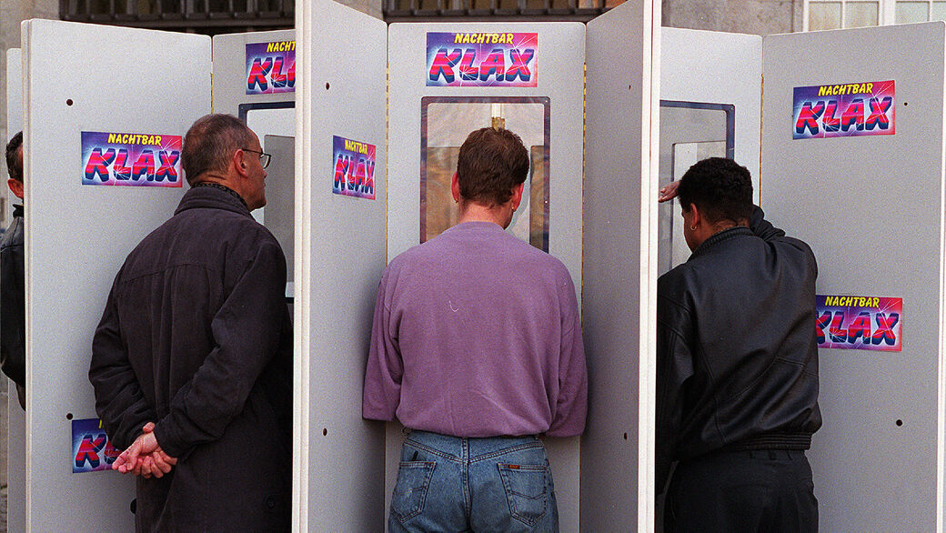 Die Männer stehen vor eine Pappwand mit Fenstern und betrachten eine nackte Frau.